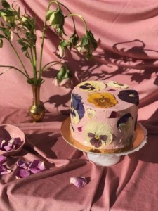 cake frosted with blush pink frosting with purple, yellow, and white pansies pressed onto it. It's styled in a pink draping fabric with a vase of flowers.