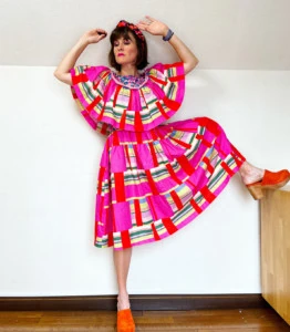 Katie wears a pink, red, green, and cream dress with red clogs. She's standing with her arms raised to demonstrate the dress bodice and sleeve flowiness.