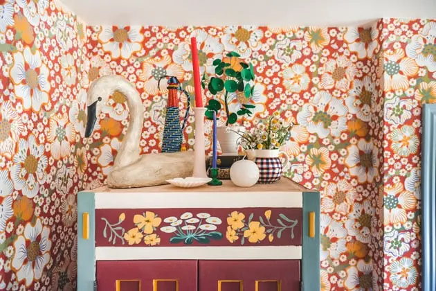 the top of a painted armoire against a red floral wallpaper background. On top of the armoire is a sculptural duck, a candle and candlestick, a paper money plant, and some cute odds and ends.