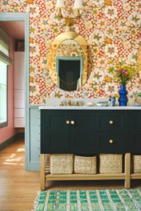 Interior shot of a green bathroom vanity with wicker baskets under it, a green rug, flowers in vases on top, and a mirror hanging above it. There's red floral wallpaper and blue painted trim.