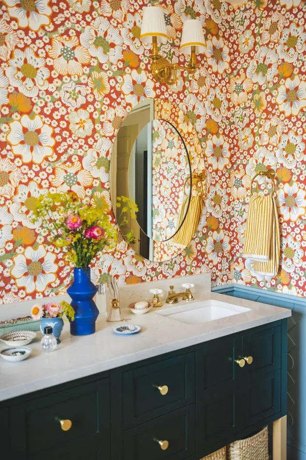 Interior shot of a bathroom with a dark emerald green vanity and red floral wallpaper. There are flowers on the vanity countertop, brass knobs and fixtures, and a blue custom wainscoting.