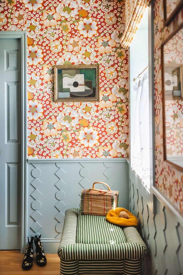 Interior shot of a bathroom. There's red floral wallpaper and framed art prints on the walls, blue textured wainscoting and trim, wooden floors, yellow window treatments, and eclectic styling.