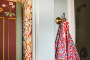 A pink and blue batik-patterned bathrobe hanging on a brushed gold hook in a bathroom. You can see a doorway and a red, floral bathroom on the side of the image.