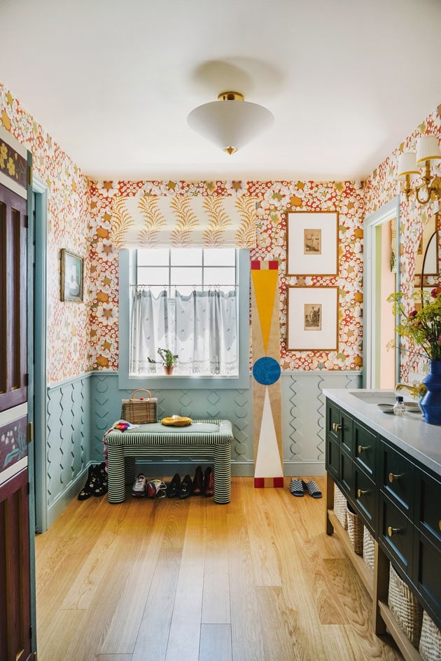 Interior shot of a bathroom. There's red floral wallpaper and framed art prints on the walls, blue textured wainscoting and trim, wooden floors, yellow window treatments, and eclectic styling.