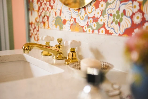 A brass faucet with white ceramic knobs on a white marble countertop. The wallpaper behind it is red and floral.