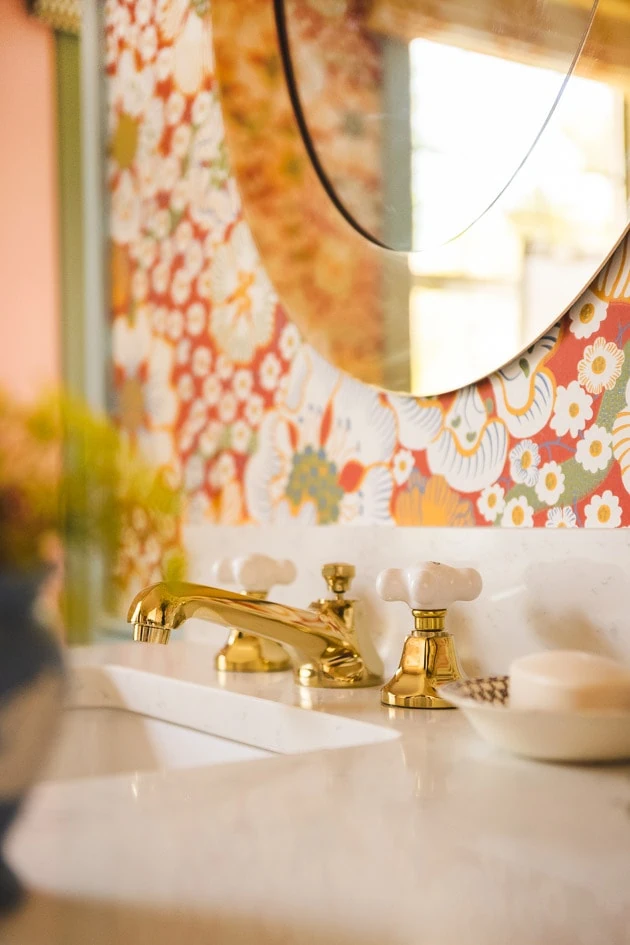 Brushed brass faucets on a marble countertop with a periwinkle vase of flowers. There's red floral wallpaper in the background.