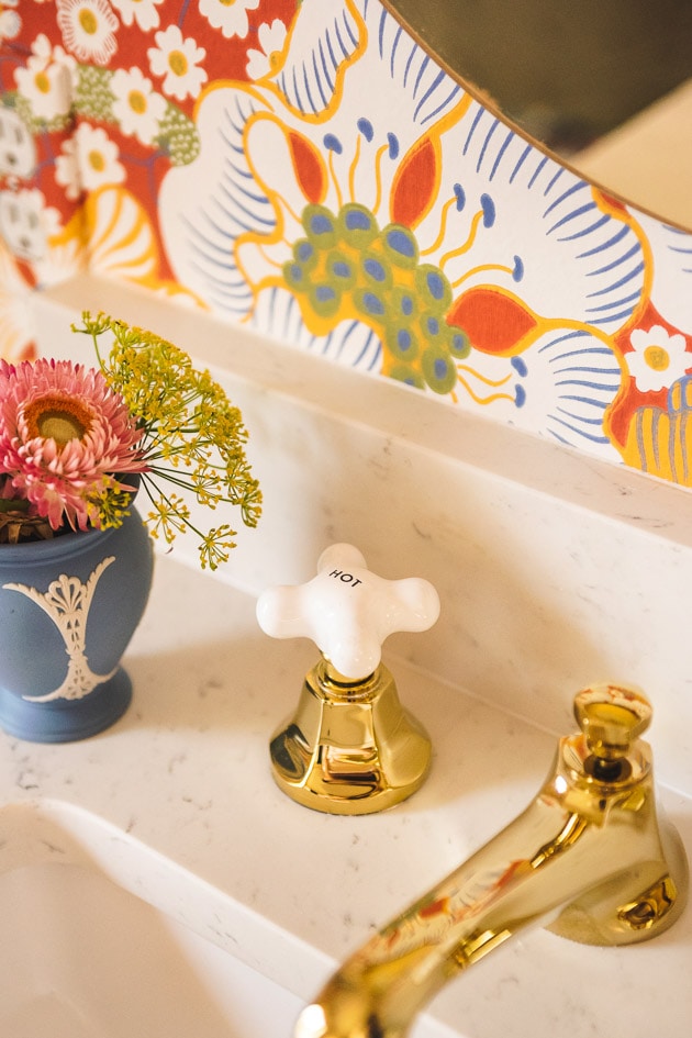 Brushed brass faucets on a marble countertop with a periwinkle vase of flowers. There's red floral wallpaper in the background.