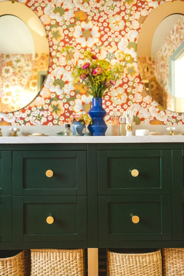Interior shot of a bathroom with a dark emerald green vanity and red floral wallpaper. There are flowers on the vanity countertop, brass knobs and fixtures, and brown wicker baskets under the vanity.