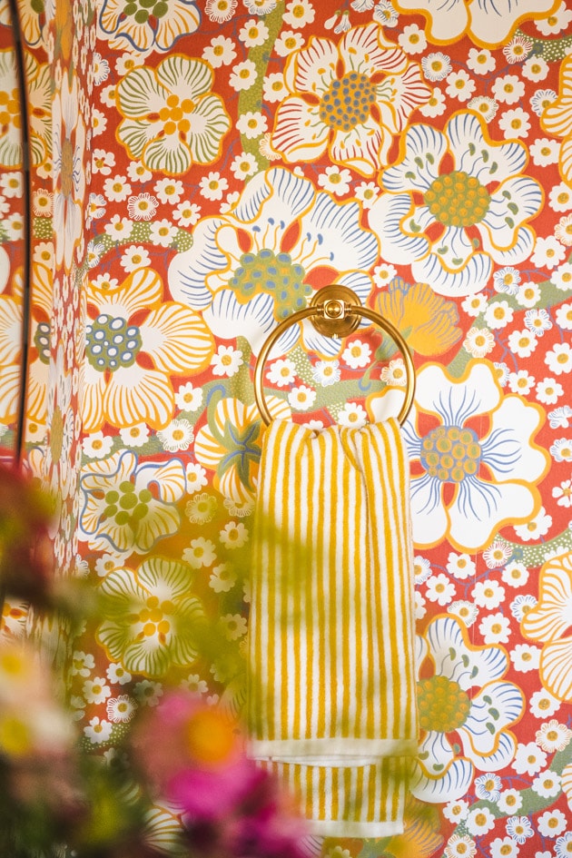 A yellow striped hand towel hangs on a brass ring on the wall. The out-of-focus silhouette of flowers in a vase shades some of the image.