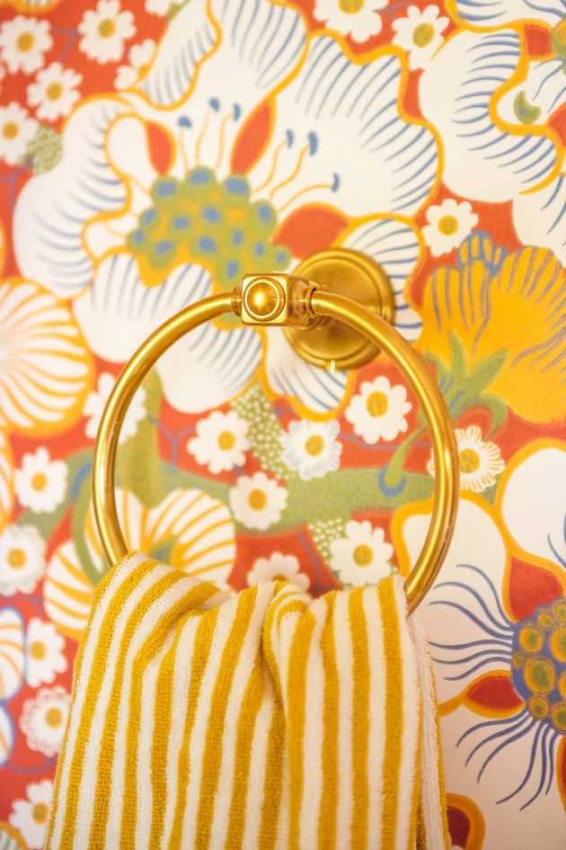 Close up of a brass towel ring. A yellow and white striped hand towel hangs from it, and there's red floral wallpaper in the back.