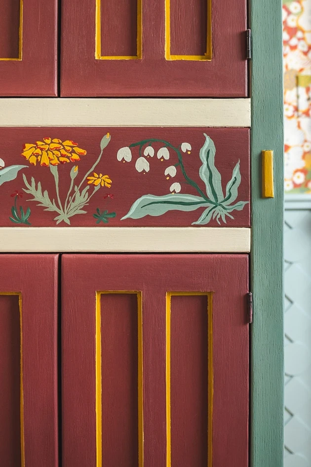 A painted cupboard. It is burgundy with green, mustard, and white accents.