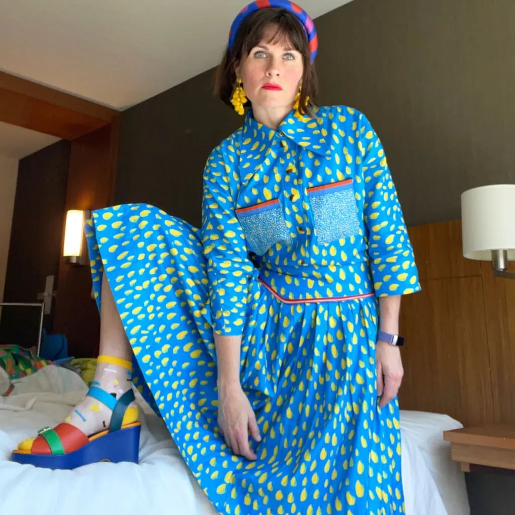 Katie wears a blue dress with painterly yellow marks and red details. Her earrings are yellow, her headband is blue with red splotches, and she's wearing blue and green wedge sandals with red and yellow socks underneath.