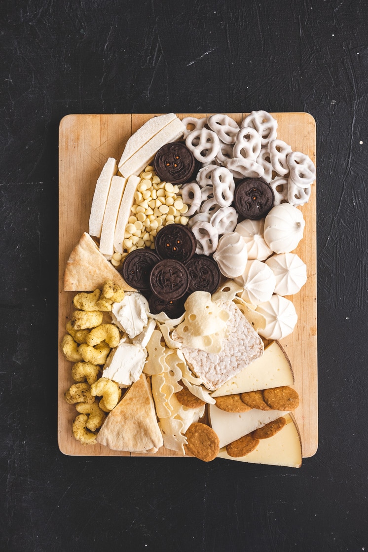 Closeup of a ghost shaped Halloween snack board. It includes oreos, yogurt-covered pretzels, merengues, jicama, white chocolate chips, pita slices, ghost-shaped potato crisps, rice puffs, cheese, goat cheese, and crackers.