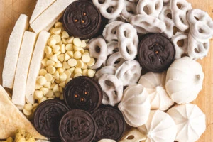 Closeup of a ghost shaped Halloween snack board. It includes oreos, yogurt-covered pretzels, merengues, jicama, and white chocolate chips.