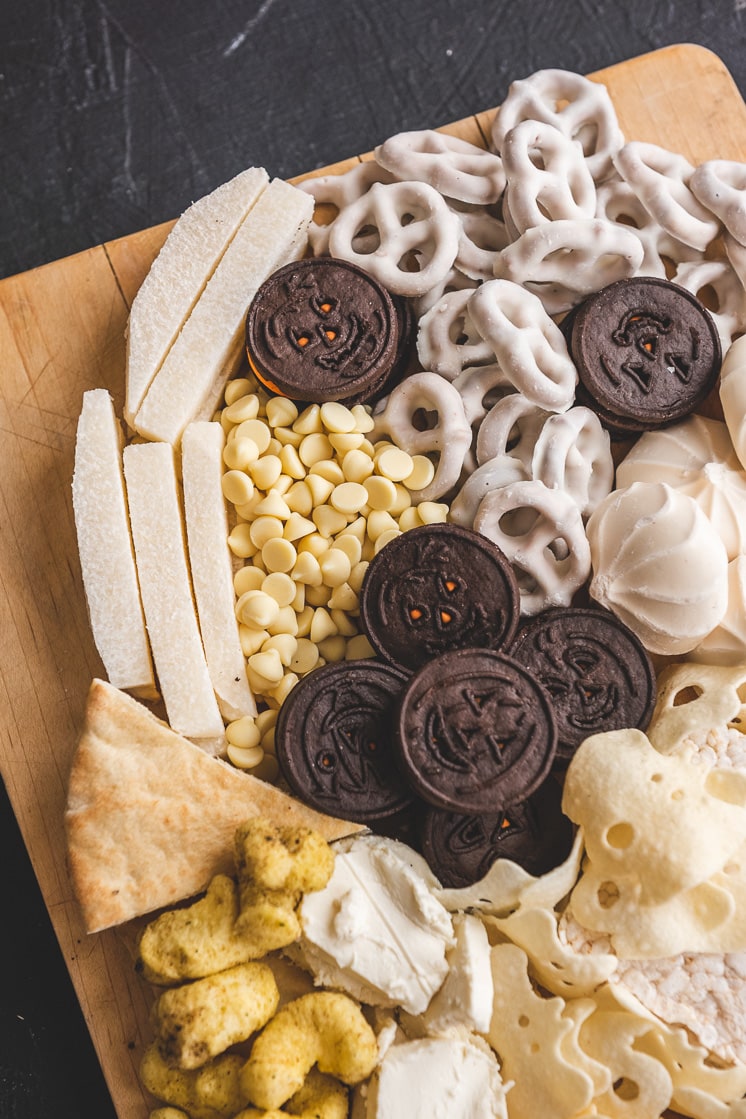 Closeup of a ghost shaped Halloween snack board. It includes oreos, yogurt-covered pretzels, merengues, jicama, white chocolate chips, pita slices, ghost-shaped potato crisps, rice puffs, cheese, goat cheese, and crackers.