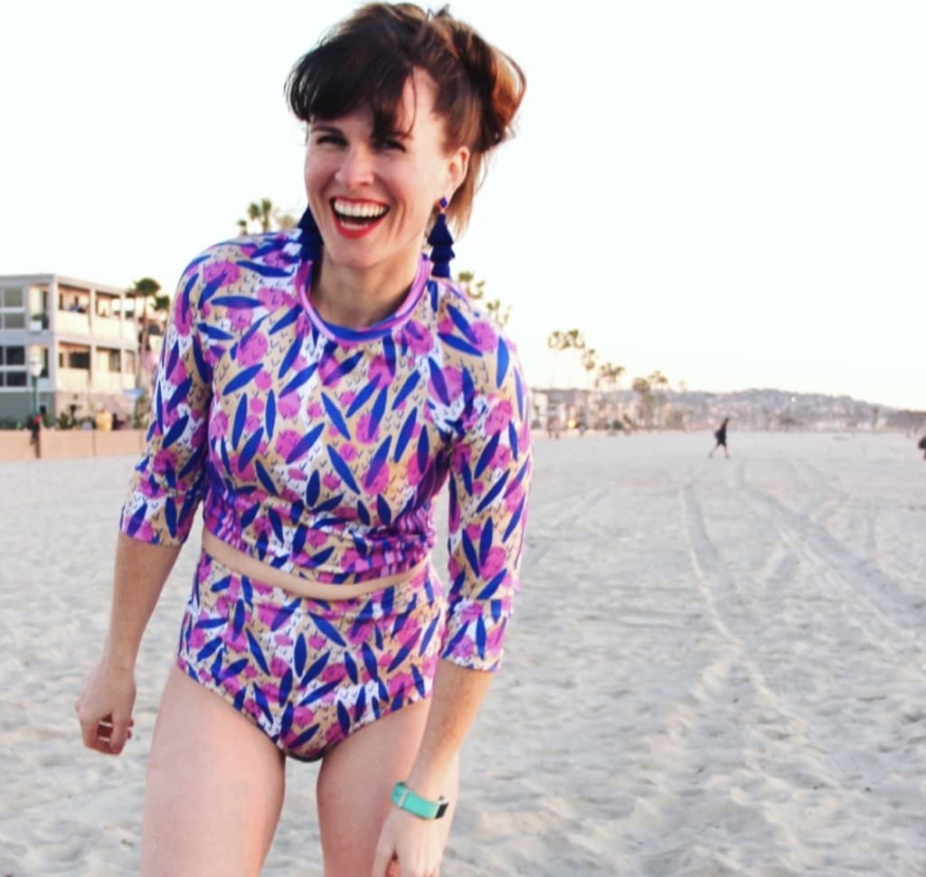 Katie on a beach wearing a pink and blue bathing suit she designed. It has two pieces, and the top is a 3/4 length sleeve tankini. The bottom is high waisted. 