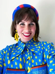 Katie Kortman wearing a blue blouse with yellow and red accents and a blue headband with red accents, smiles at the camera.