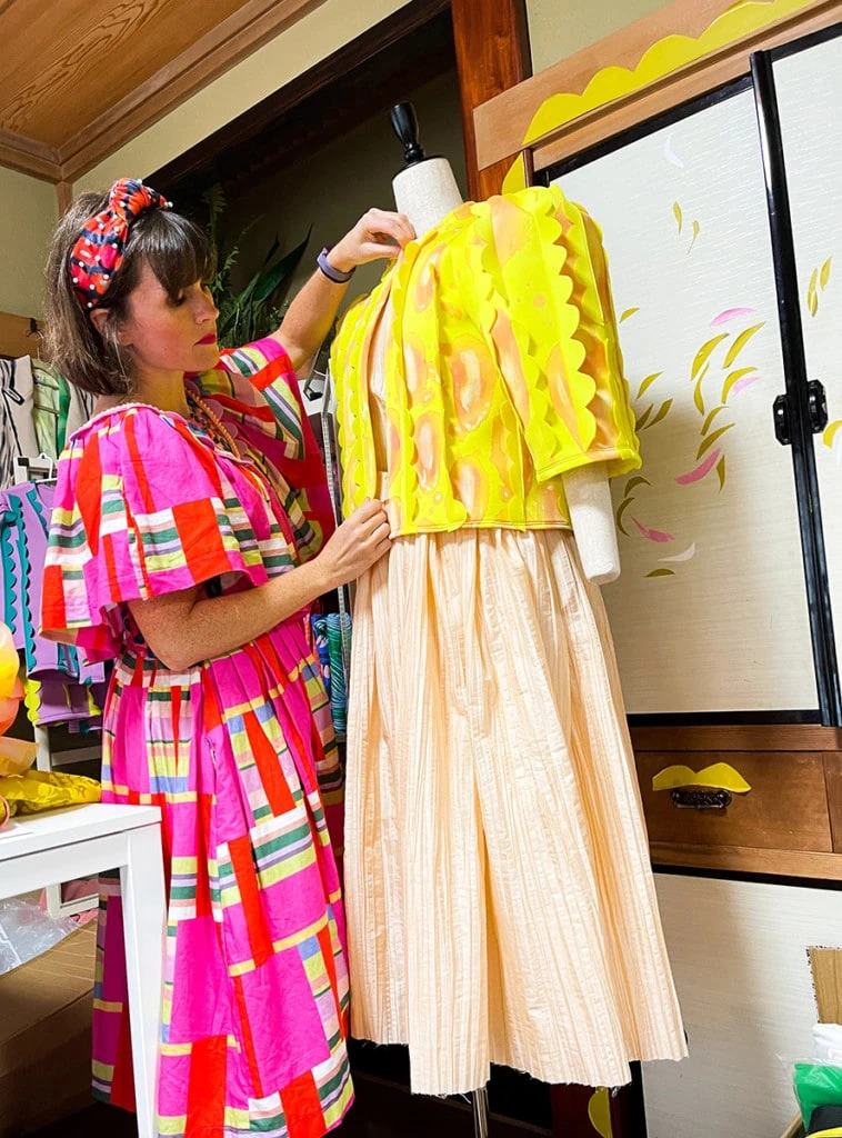 Katie works on a yellow jacket while standing by a dress model. Katie's wearing a pink, red, yellow, and green colorful dress.
