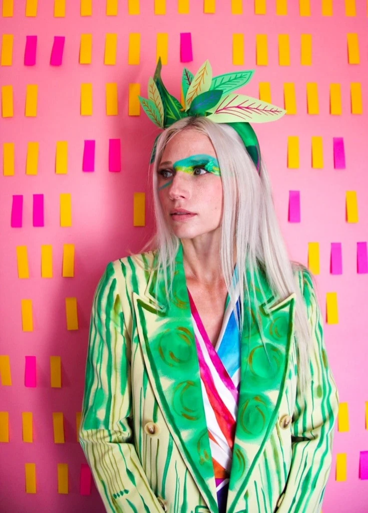 A model wearing a green Katie Kortman blazer with a pink, white, and blue top underneath. She has a headdress made of fabric leaves and she's standing by a pink and yellow wall. 