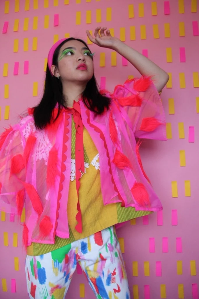 A model wears Katie's pink overjacket with a yellow top underneath and white pants with colorful details. The backdrop is pink with yellow and pink rectangles.