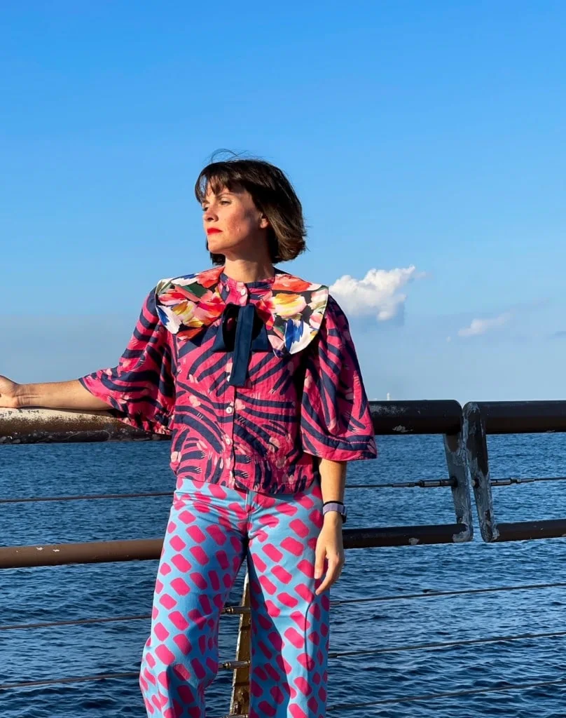 Katie Kortman wearing bright prink and blue pands and a blouse standing by the ocean.