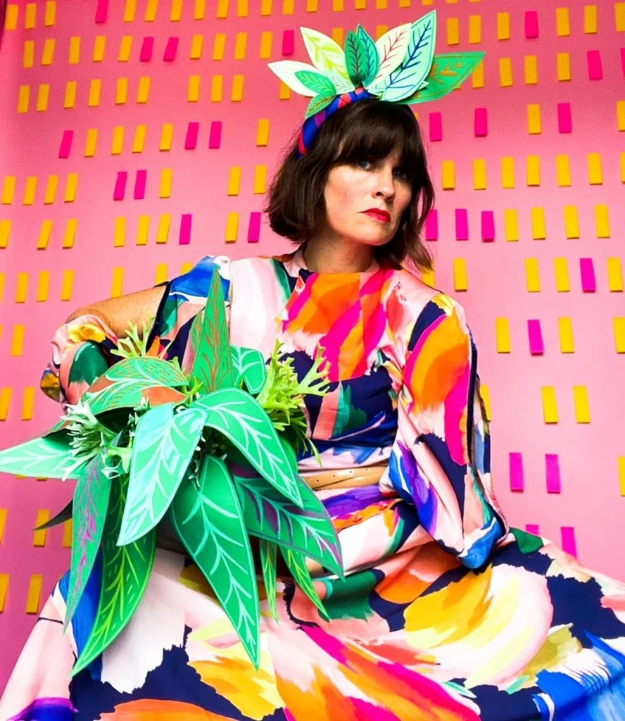 Katie Kortman modeling a vibrant dress and holding fabric plants in front of a pink wall