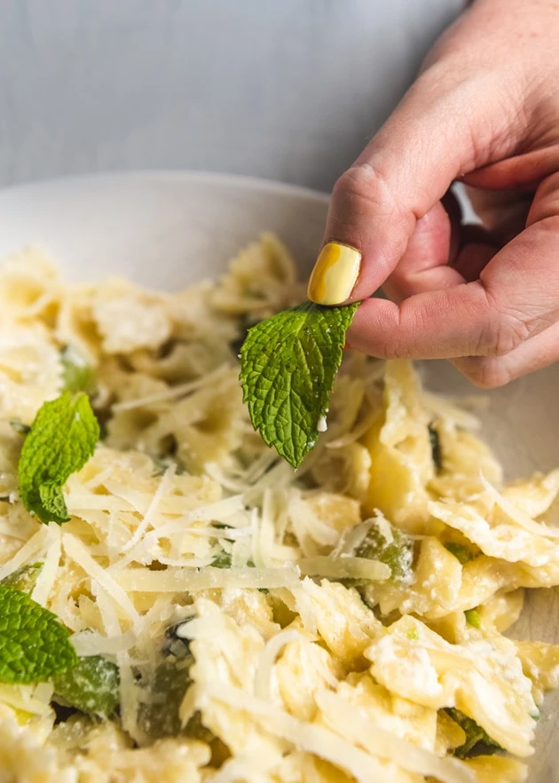 Brittany puts a mint leaf into a bowl of bowtie pasta with peas and cheese. She has a yellow-painted thumbnail.