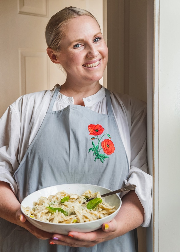 Brittany stands in a doorway holding a plate of bowtie pasta with cheese and peas. She's smiling and wearing a white blouse, as well as a blue apron with red poppies on it.