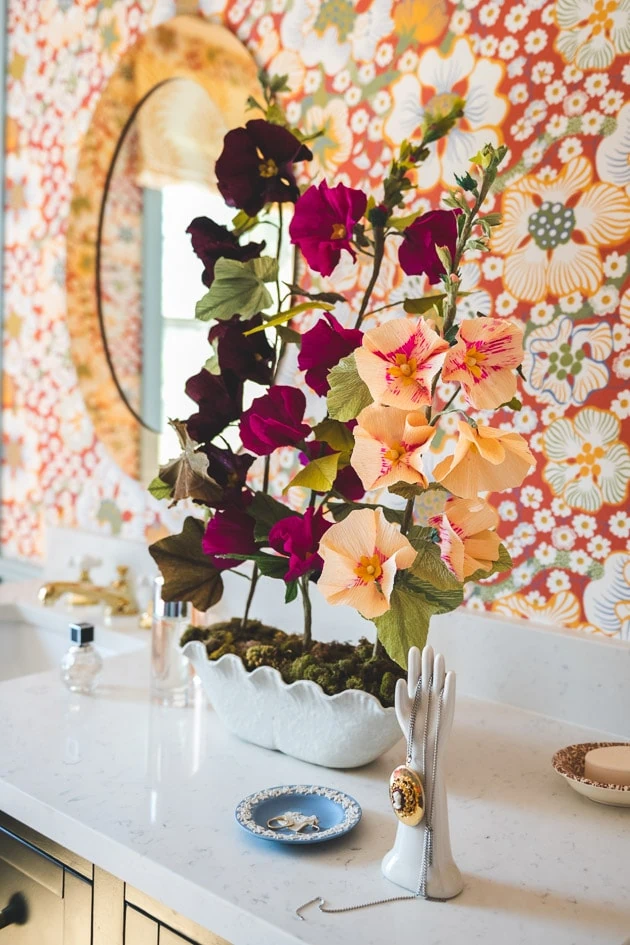 paper hollyhocks on a bathroom counter among ceramic odds and ends with a mirror and red floral wallpaper in the background.