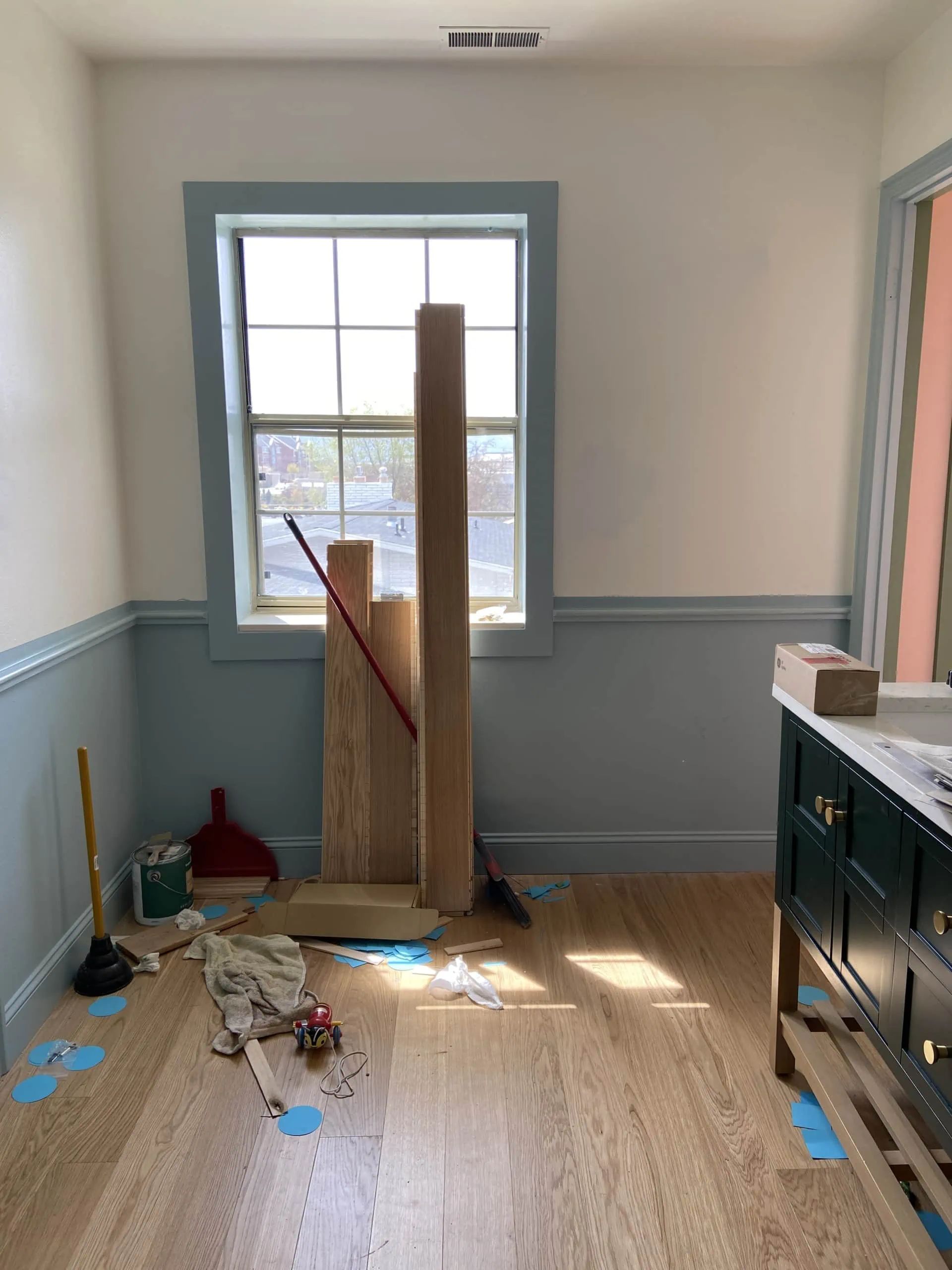 Interior shot of a bathroom with wooden floors and blue and white walls. There's blue-painted trim at waist-height around the room, and some boards and materials are cluttered in the corner and under a window.