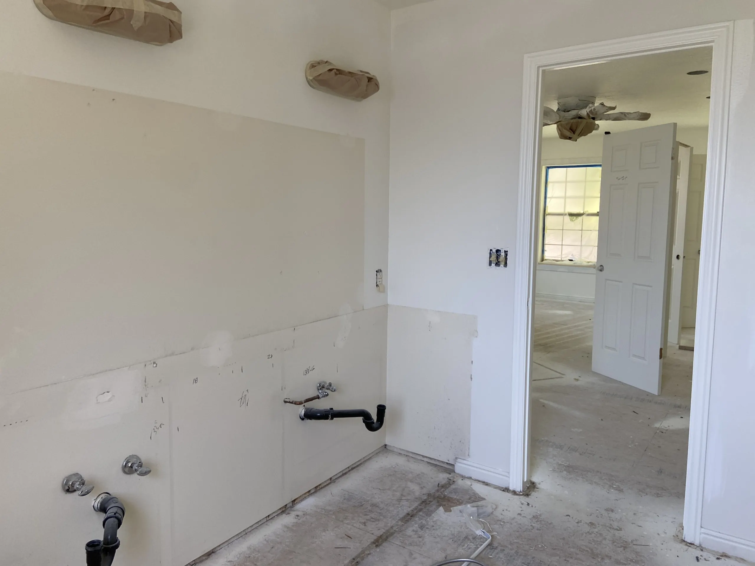a blank, unfinished room with sheetrock walls and a dusty subfloor. There's a doorway that leads to another unfinished space in the image. One of the walls has mysterious plumbing coming out of it.